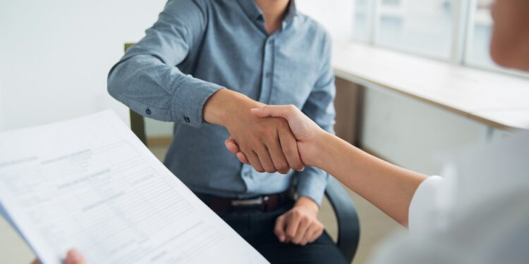 Smiling Asian businessman wearing shirt sitting in office and shaking hand of female partner. Woman holding document