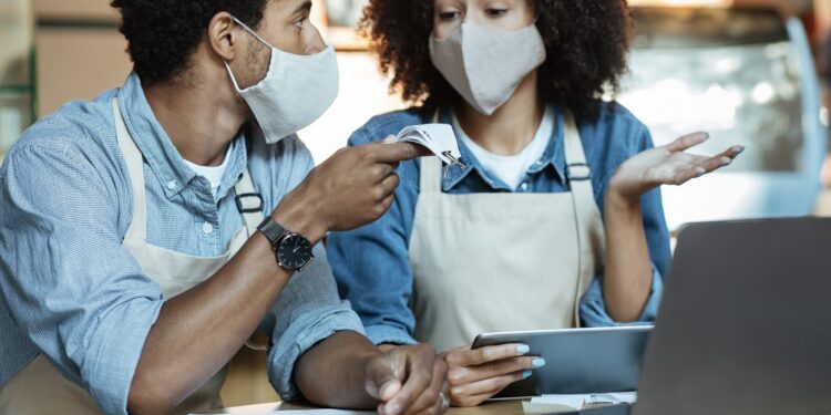 Financial problems in family small business during covid-19 pandemic. Sad young african american couple in aprons and protective masks gesturing and talking about bills and work with laptop in cafe