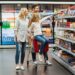 Cheery little girl sitting on a shopping cart and choosing candy with her parents at the supermarket