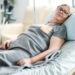 Elderly age woman laying on a bed in a nursing home covered with a blanket.