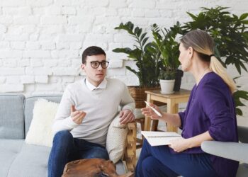 Picture of frustrated young Caucasian man wearing sweater and eyeglasses sitting on comfortable couch, sharing his personal problems with middle aged female counselor during therapy session