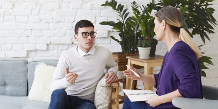 Picture of frustrated young Caucasian man wearing sweater and eyeglasses sitting on comfortable couch, sharing his personal problems with middle aged female counselor during therapy session