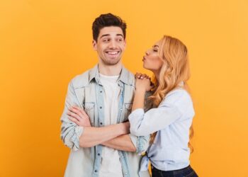 Portrait of young lovely people in basic clothing expressing love and affection while woman kissing man on cheek isolated over yellow background