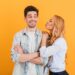 Portrait of young lovely people in basic clothing expressing love and affection while woman kissing man on cheek isolated over yellow background