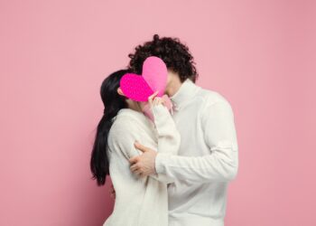 Young, beautiful couple in love on pink studio background. Saint Valentine's Day, love, relationship and human emotions concept. Copyspace. Young man and woman hiding behind the heart shaped card.