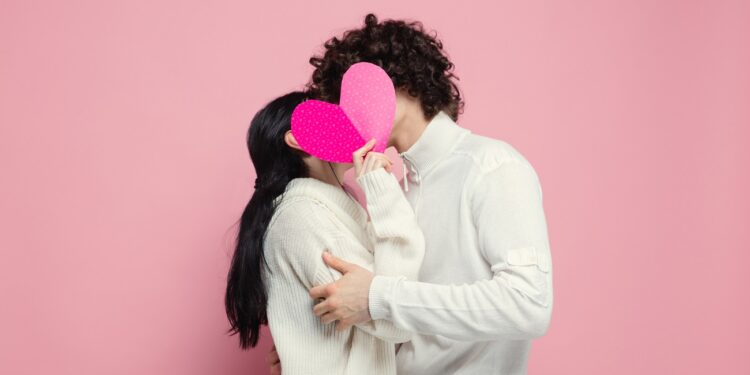 Young, beautiful couple in love on pink studio background. Saint Valentine's Day, love, relationship and human emotions concept. Copyspace. Young man and woman hiding behind the heart shaped card.