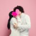 Young, beautiful couple in love on pink studio background. Saint Valentine's Day, love, relationship and human emotions concept. Copyspace. Young man and woman hiding behind the heart shaped card.