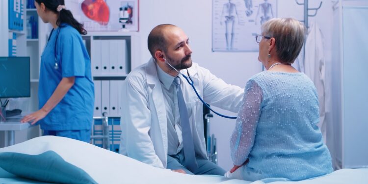 Elderly woman doctor appointment in modern private clinic while she sitts on hospital bed and the physician is listening her heart beat. Health care medicine treatment, specialist consultation