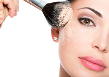 Closeup portrait of a woman applying dry cosmetic tonal foundation on the face using makeup brush.