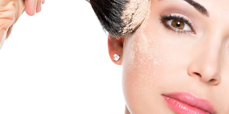 Closeup portrait of a woman applying dry cosmetic tonal foundation on the face using makeup brush.