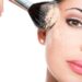 Closeup portrait of a woman applying dry cosmetic tonal foundation on the face using makeup brush.