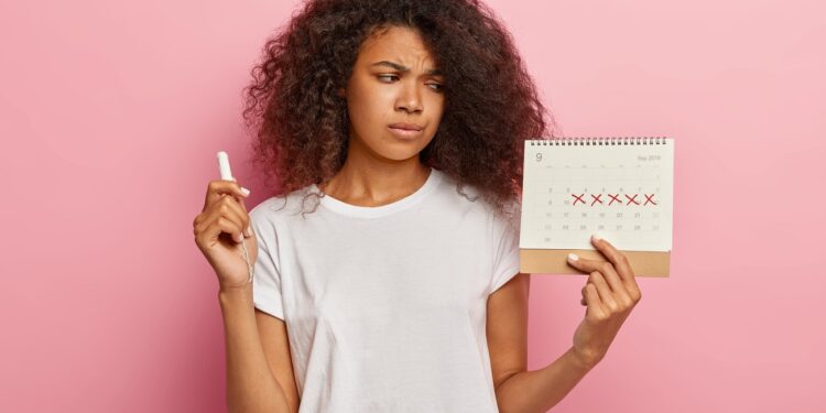 Displeased stressful dark skinned woman looks at periods calendar with marked red crosses, has painful menstruation, holds tampon for good protection. People, women health and hygiene concept