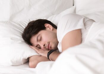 Man is asleep in cozy white bed at night, handsome young guy sleeping at home with wearable electronic device smart watch on his wrist for sleep tracking, monitoring heart rate for healthcare