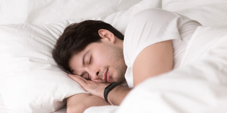 Man is asleep in cozy white bed at night, handsome young guy sleeping at home with wearable electronic device smart watch on his wrist for sleep tracking, monitoring heart rate for healthcare