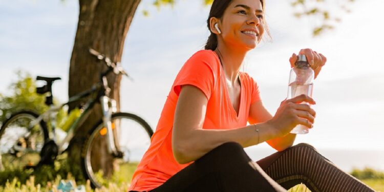 smiling beautiful woman drinking water in bottle doing sports in morning in park nature doing yoga on mat, pink fitness outfit, happy healthy lifestyle, music in earphones, bicycle on background