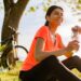smiling beautiful woman drinking water in bottle doing sports in morning in park nature doing yoga on mat, pink fitness outfit, happy healthy lifestyle, music in earphones, bicycle on background
