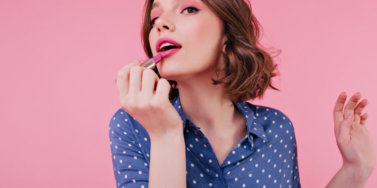 Graceful woman with tattoo doing lips makeup on bright background. Indoor portrait of magnificent short-haired girl posing with lipstick.