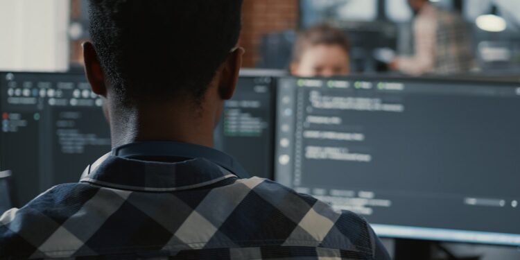 Portrait of african american programer online cloud computing on laptop sitting at desk with multiple screens parsing code. Over shoulder view of app developer working on user interface.