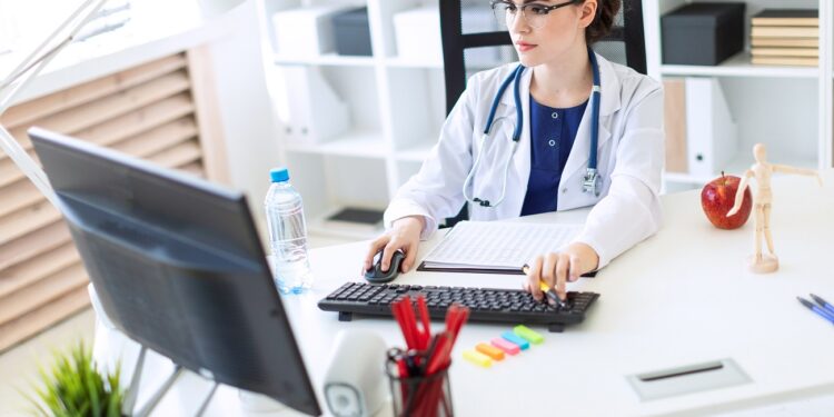 A charming young girl with glasses, a blue blouse and a white robe is sitting in the office. next to the girl an apple and a bottle of water. photo with depth of field.