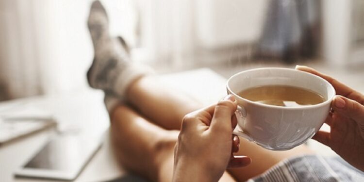 Cup of tea and chill. Woman lying on couch, holding legs on coffee table, drinking hot coffee and enjoying morning, being in dreamy and relaxed mood. Girl in oversized shirt takes break at home.