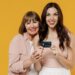 Two caucasian young smiling happy daughter mother together couple women in casual clothes hug hold in hand credit bank card isolated on plain yellow color background studio. Family lifestyle concept