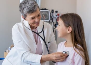 Happy little girl at the doctor for a checkup. Friendly pediatrician examining girl with stethoscope in her office. Happy senior woman auscultate the heartbeat of the child.