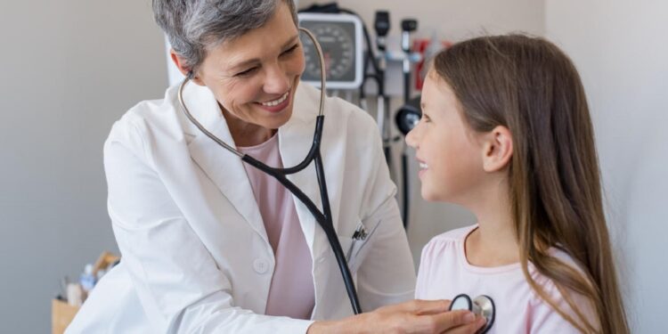 Happy little girl at the doctor for a checkup. Friendly pediatrician examining girl with stethoscope in her office. Happy senior woman auscultate the heartbeat of the child.