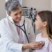 Happy little girl at the doctor for a checkup. Friendly pediatrician examining girl with stethoscope in her office. Happy senior woman auscultate the heartbeat of the child.