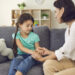 Young mother sitting on sofa with daughter, holding her hands and talking to her seriously at home with room interior at background. Solving problems in children education concept