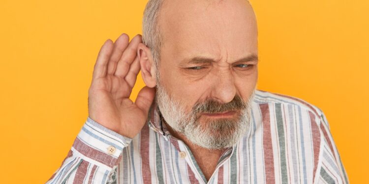 Portrait of frowning frustrated bearded male pensioner in striped shirt keeping hand at his ear, listening attentively, trying to hear indistinct talking. Hearing problems and eavesdropping