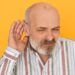 Portrait of frowning frustrated bearded male pensioner in striped shirt keeping hand at his ear, listening attentively, trying to hear indistinct talking. Hearing problems and eavesdropping
