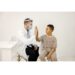 Male doctor sitting at table and wearing face mask. Man wearing white medical gown while isolated on white backgroun. Man give a high five to a little boy.