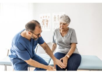 Photo of senior woman having some knee pain. She's at doctor's office having medical examination by a male doctor. The doctor is touching the sensitive area and trying to determine the cause of pain.
