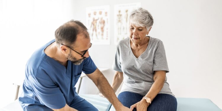 Photo of senior woman having some knee pain. She's at doctor's office having medical examination by a male doctor. The doctor is touching the sensitive area and trying to determine the cause of pain.