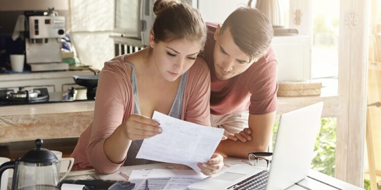 Young couple managing finances, reviewing their bank accounts using laptop computer and calculator at modern kitchen. Woman and man doing paperwork together, paying taxes online on notebook pc
