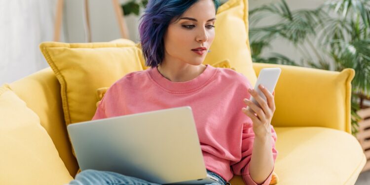 Freelancer with colorful hair and laptop looking at smartphone and lying on sofa