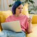 Freelancer with colorful hair and laptop looking at smartphone and lying on sofa