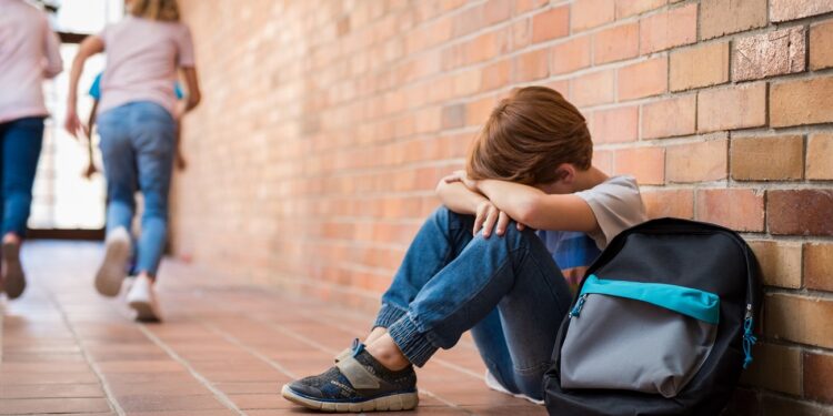 Little boy sitting alone on floor after suffering an act of bullying while children run in the background. Sad young schoolboy sitting on corridor with hands on knees and head between his legs.