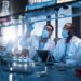 Modern Medical Research Laboratory: Two Scientists Wearing Face Masks use Microscope, Analyse Sample in Petri Dish, Discuss Innovative Technology. Advanced Scientific Lab for Medicine, Biotechnology