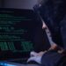 Computer hacker in hooded shirt typing software algorithms and stealing data from computer network on laptop.