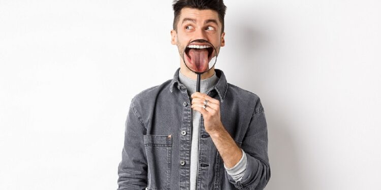 Handsome positive guy showing white perfect teeth and tongue with magnifying glass, looking left at logo, standing against white background.