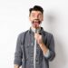 Handsome positive guy showing white perfect teeth and tongue with magnifying glass, looking left at logo, standing against white background.