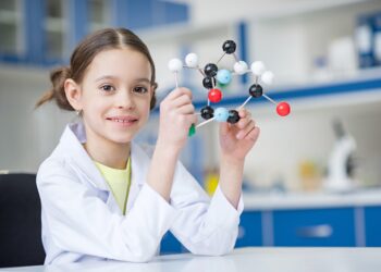 Girl scientist in lab coat holding molecular model and smiling at camera; Shutterstock ID 601069865; purchase_order: -; job: -; client: -; other: -