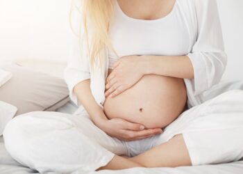 Young pregnant lady touching her belly sitting in her bedroom in afternoon. Pregnancy concept.