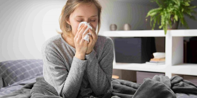 Sick young Caucasian woman covered with grey blanket sitting on bed with closed eyes, blowing nose with napkin. Illness, pain concept