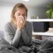 Sick young Caucasian woman covered with grey blanket sitting on bed with closed eyes, blowing nose with napkin. Illness, pain concept
