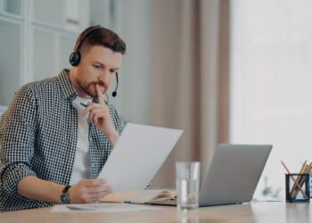 Young focused bearded business professional in casual wear and headset while working with laptop and documents during online meeting with colleagues. Remote work at home