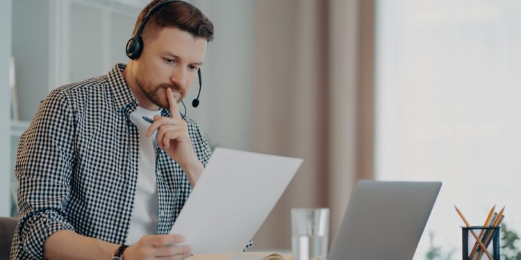 Young focused bearded business professional in casual wear and headset while working with laptop and documents during online meeting with colleagues. Remote work at home