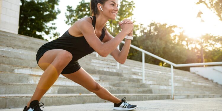Portrait of a smiling fitness woman in earphones doing stretching exercises on stairs outdoors