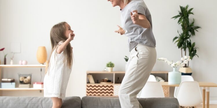 Cute little girl having fun playing with smiling grandmother jumping on couch together, happy granny and active kid grandchild dancing on sofa, grandma and granddaughter laughing playing at home